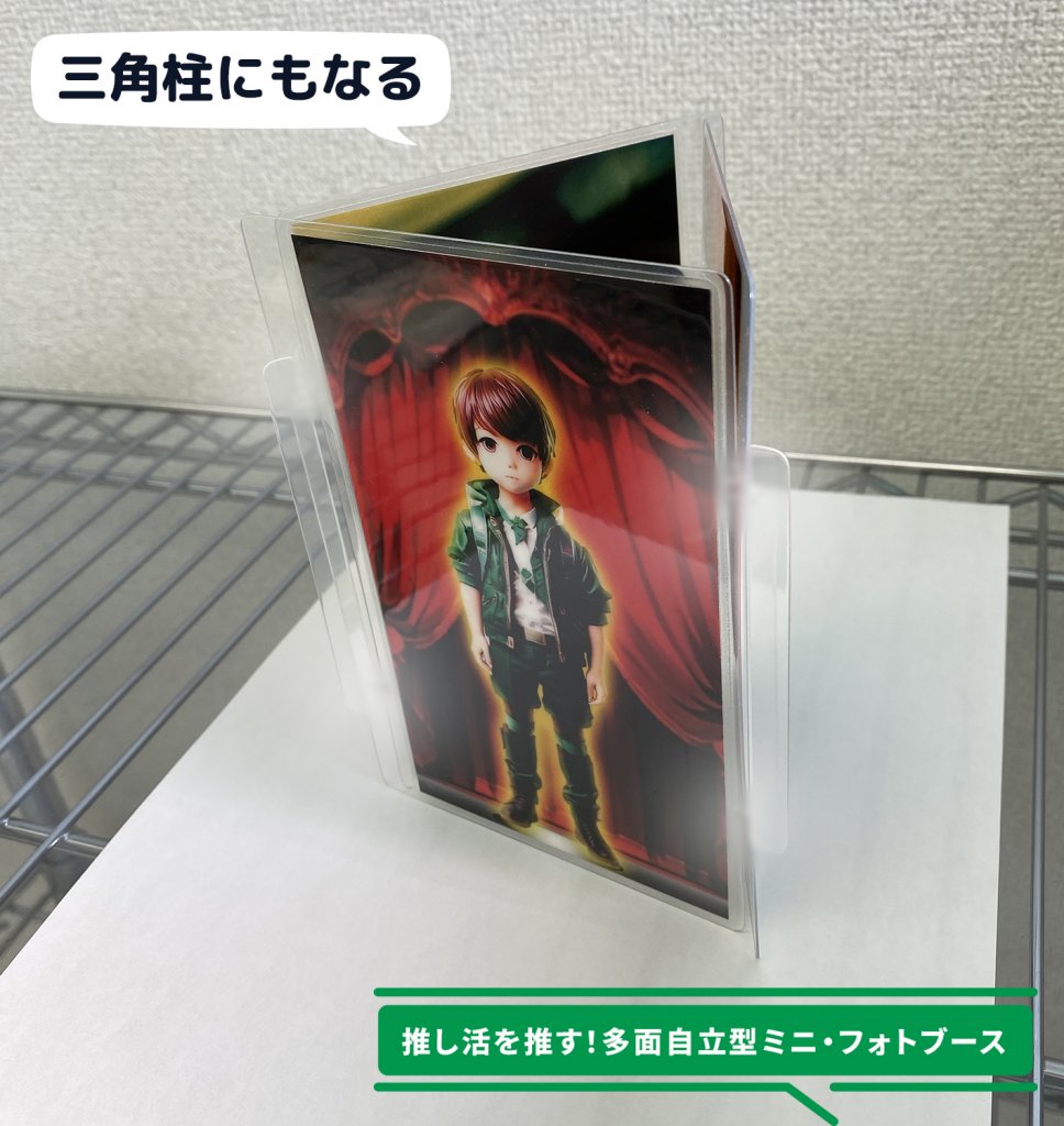 金属製の棚の上に、アニメ風の男性キャラクターのイラストが描かれた三連のミニフォトブースプレートが、三角形の柱状に組み立てられて置かれている様子。『三角柱にもなる』という吹き出しがあり、このミニフォトブースが三角柱の形状でも使用できることが示されている