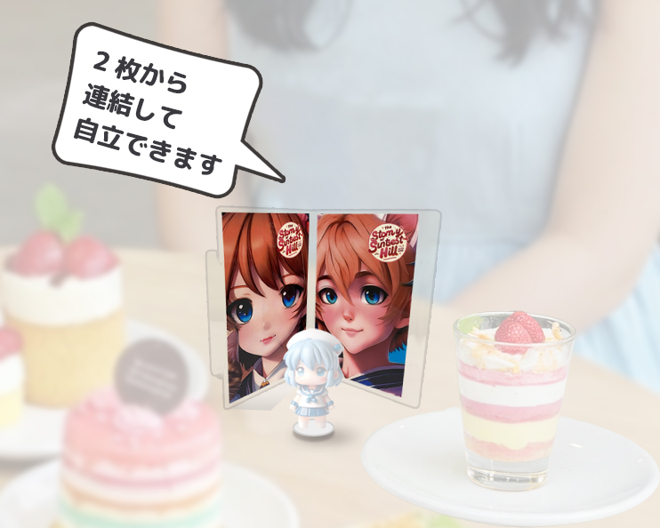 テーブルの上に、アニメ風の少女のイラストが描かれた連結式のミニフォトブース、白いワンピースを着た小さなフィギュア、そして2種類のデザートが置かれた、可愛らしい雰囲気の写真