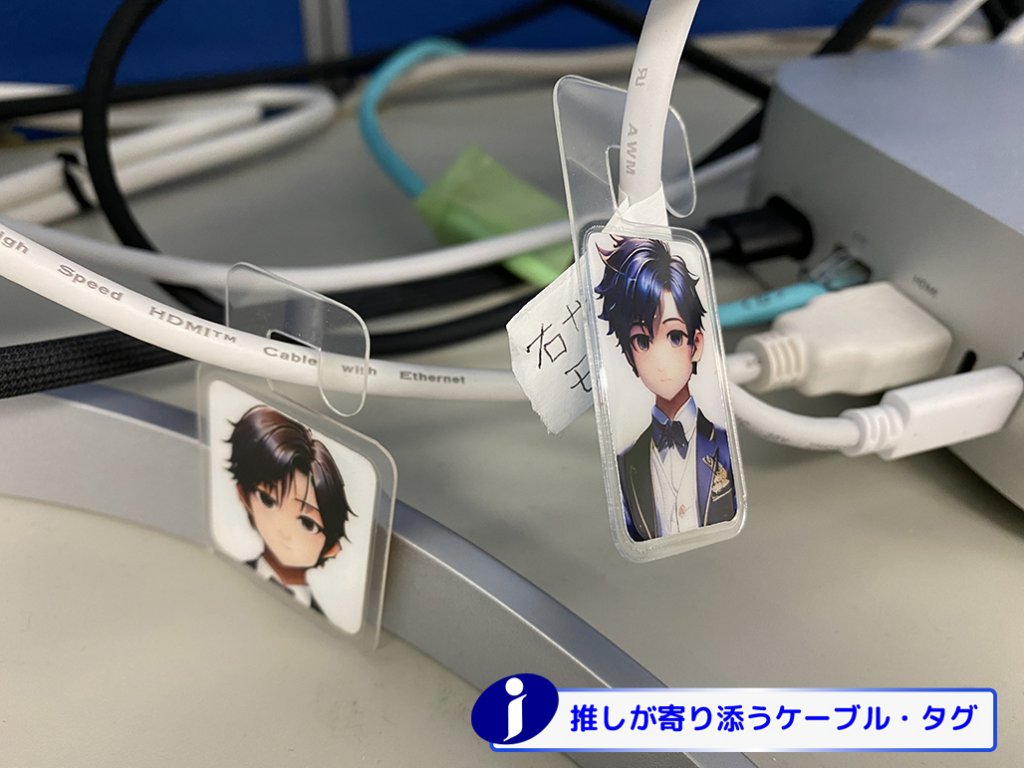 デスク上の白いケーブルに、アニメキャラクターのイラストが入った透明なケーブルタグが取り付けられている。