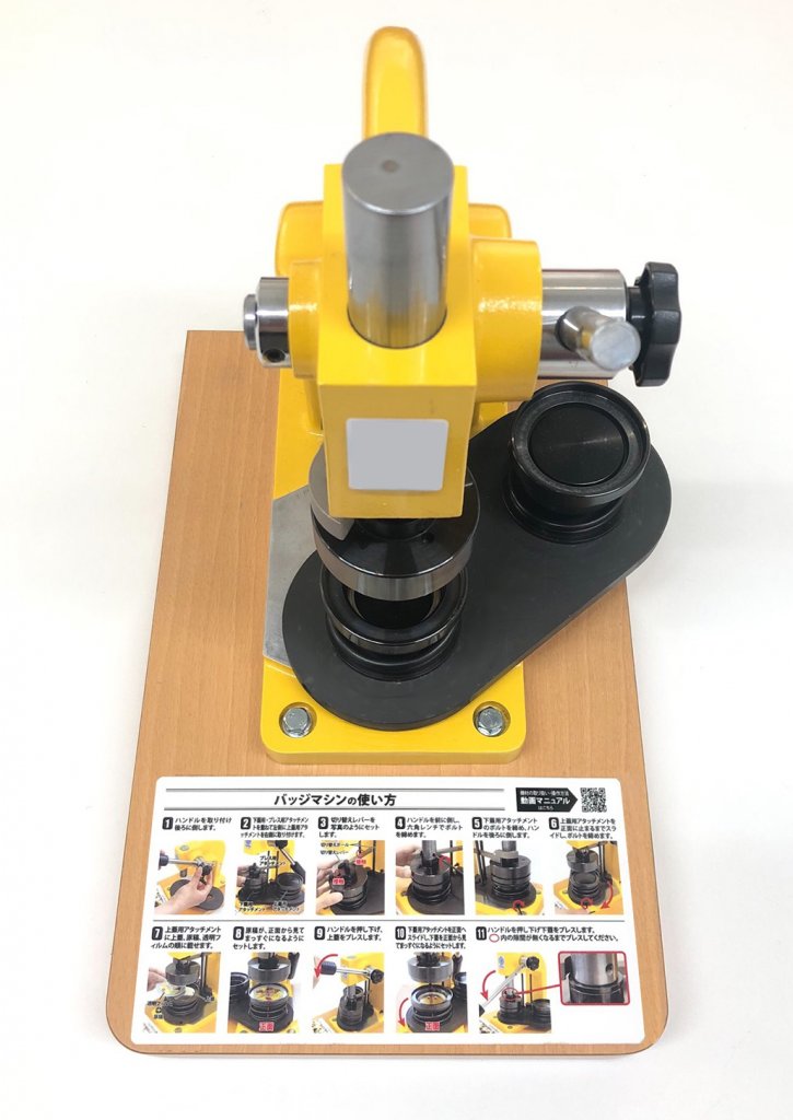 黄色と黒のバッジマシンが木製の台座に設置された上からのクローズアップ画像。台座には、バッジマシンの使い方を説明した写真入りの手順書が貼られている。