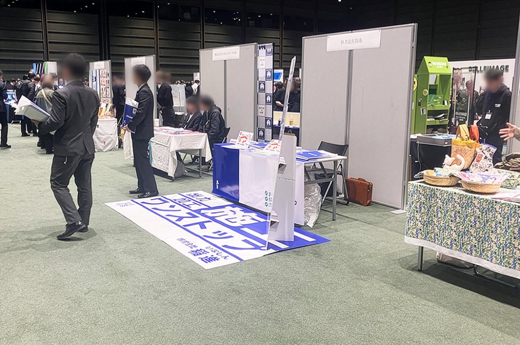 展示会場のブースの様子。手前には「企画・施工・ワンストップ」と大きく書かれた青と白のフロアマットが敷かれ、奥には説明員と来場者が談笑している複数の企業ブースが並んでいる。